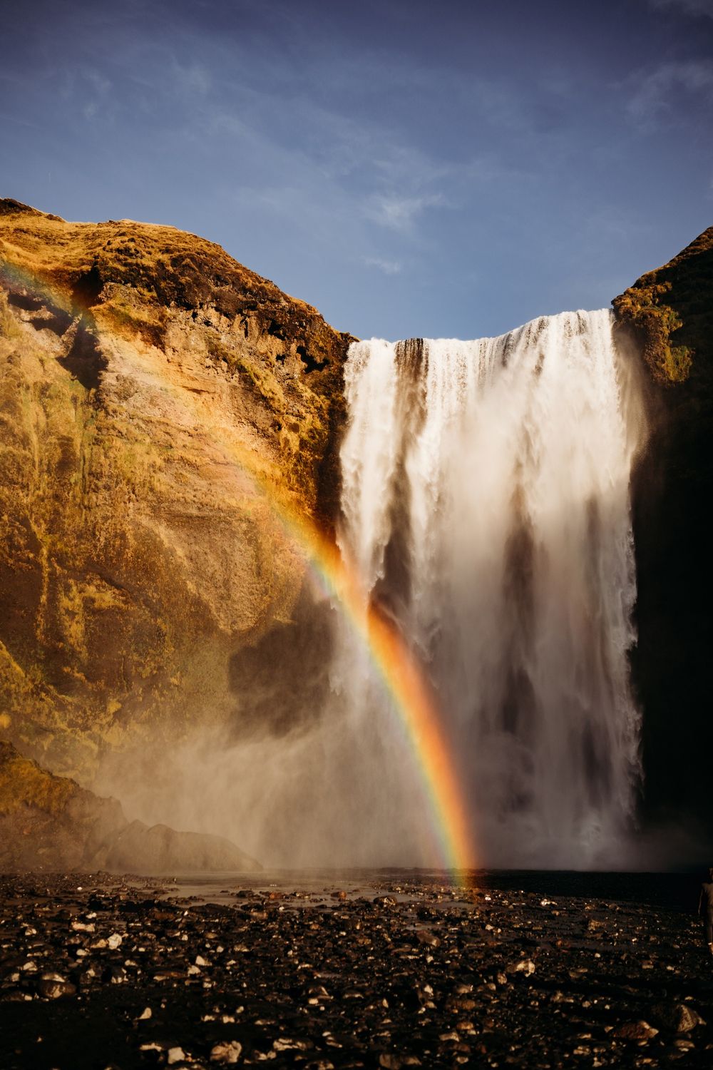 Skogafoss waterfall in Iceland with a vivid rainbow arching at its base, framed by golden cliffs and a blue sky.