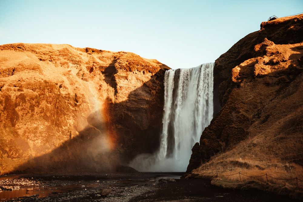 A stunning waterfall cascades between golden rocky cliffs with a rainbow in the mist under a clear blue sky.
