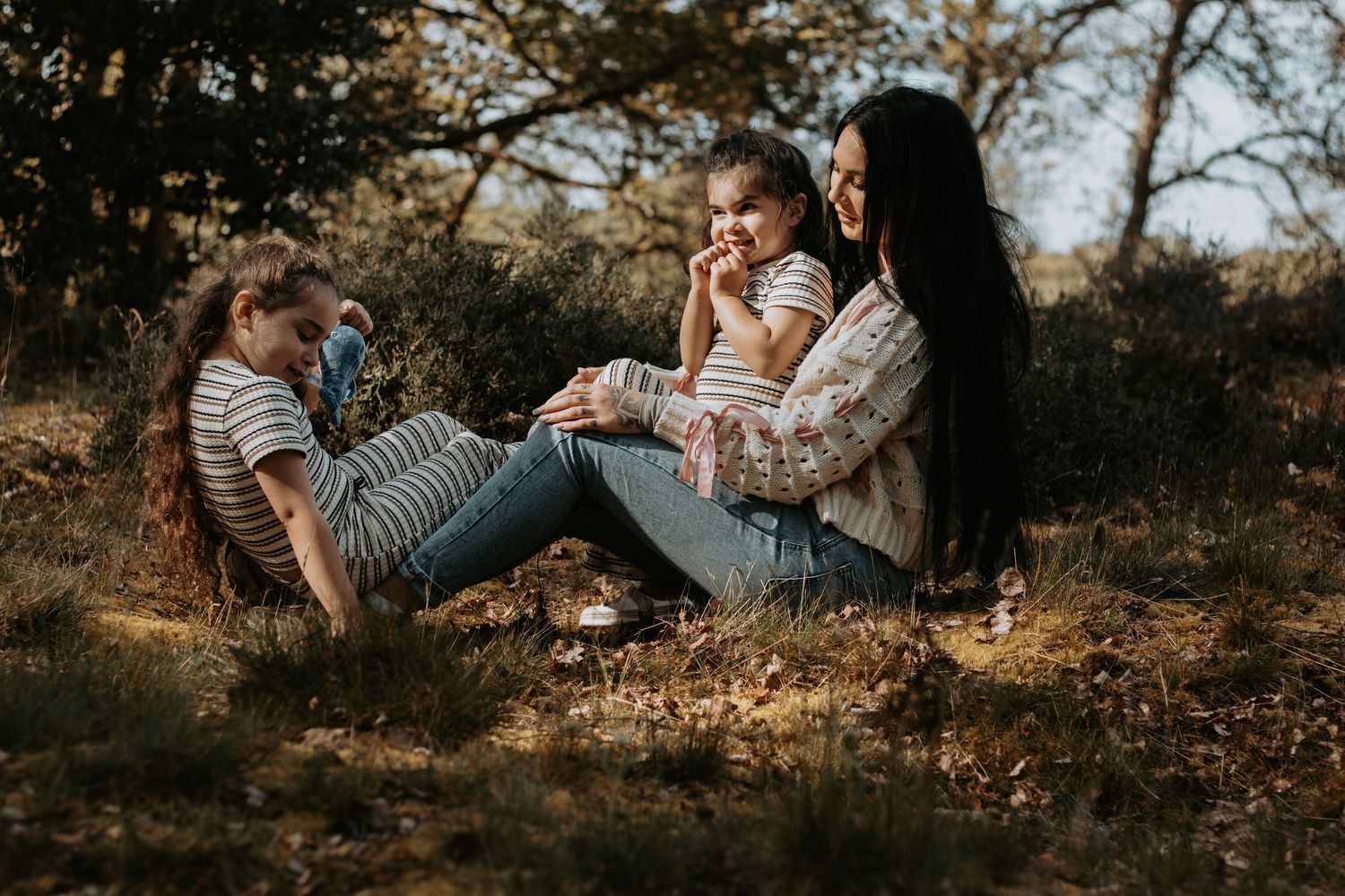 A mother sits outdoors with her two daughters, laughing and enjoying a peaceful picnic in a sunlit forest setting.