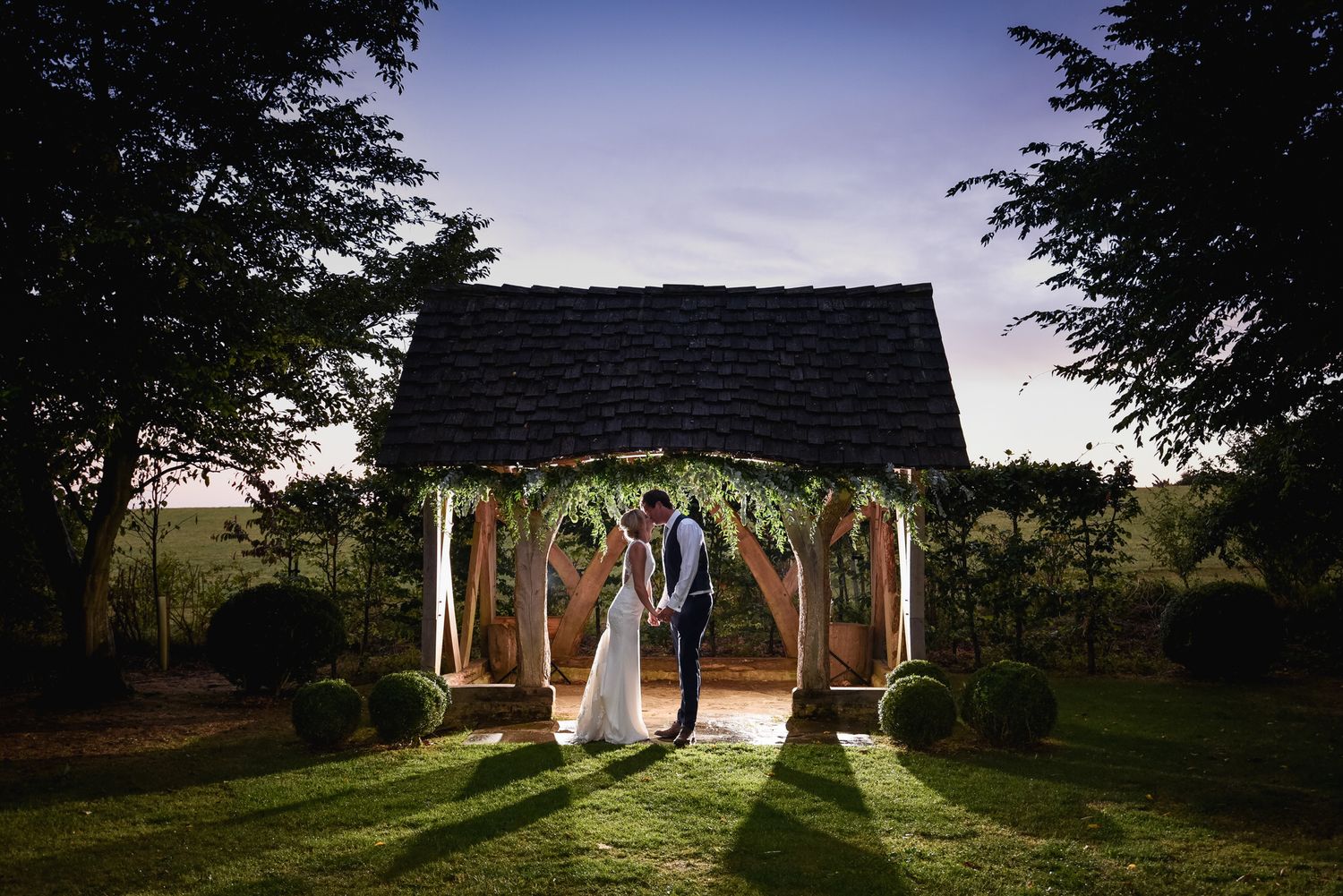 Bride and groom holding hands under a wooden gazebo adorned with greenery at dusk in a lush garden setting at Cripps Barn Wedding Venue