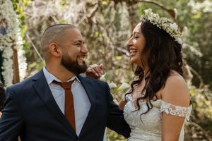 Smiling newlywed couple laughing together outdoors, bride wearing floral crown and lace gown.