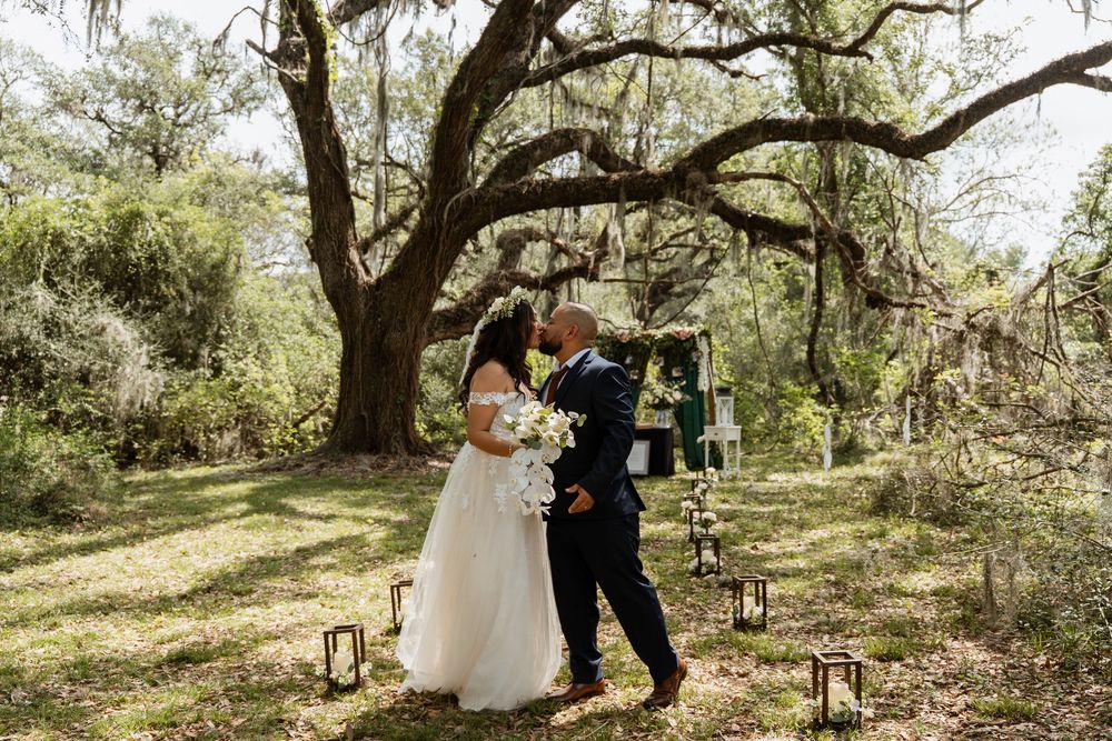 Bride and groom share first kiss under a large oak tree at outdoor woodland wedding.