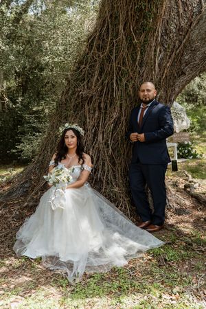 Bride in white gown and groom in navy suit posing by a large tree in a forest setting.