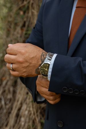 Groom in navy suit adjusting silver watch on wrist with wedding ring visible outdoors.