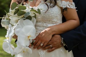 Close-up of bride and groom hands showing wedding rings with white orchid bouquet.