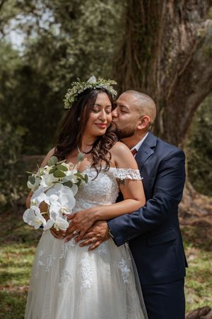Groom in navy suit kissing bride wearing floral crown holding white orchid bouquet outdoors.
