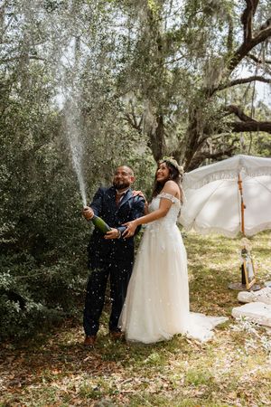 Joyful bride and groom popping champagne bottle outdoors under Spanish moss-draped trees.
