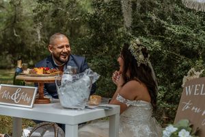 Bride and groom laughing together at their rustic outdoor wedding reception table in the woods.