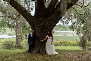 Bride and groom leaning against a massive oak tree with a scenic waterway in the background.