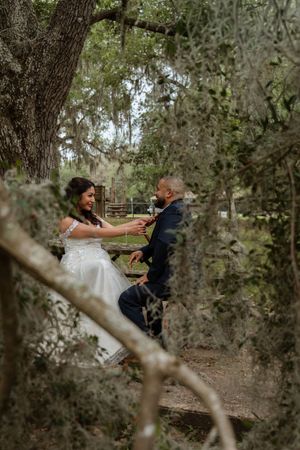 Bride and groom share a romantic moment on a bench surrounded by moss-draped trees in a lush forest setting.