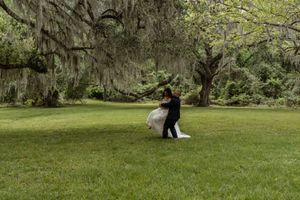 Groom twirls bride in white wedding gown on a green lawn beneath sprawling moss-covered oak trees.