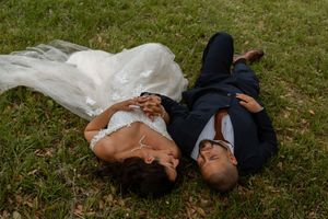 Bride and groom lay together on green grass, holding hands and gazing at each other on their wedding day.