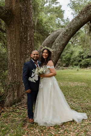 Smiling bride and groom pose together beneath a large oak tree, bride holding white floral bouquet with flower crown.