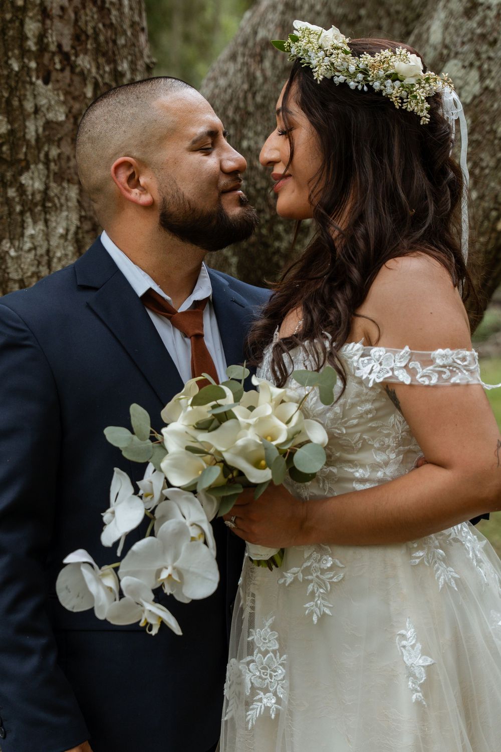 Bride and groom share an intimate moment, bride holding white orchid bouquet and wearing a floral crown in nature.