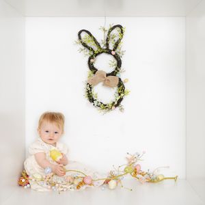 Baby girl in white dress sits beside Easter floral garland beneath a decorative bunny wreath with bow.