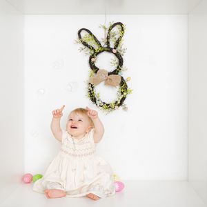 A smiling baby in a white dress sits on the floor pointing up at an Easter bunny wreath with colorful eggs nearby.