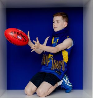 Young boy in AFL West Coast Eagles jersey kneeling and catching a red Sherrin football in a blue studio setting.