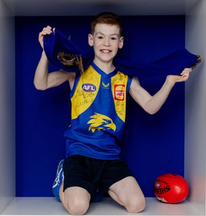 Young AFL fan kneeling in West Coast Eagles blue and yellow jersey, holding team scarf and smiling, with a red football nearby.