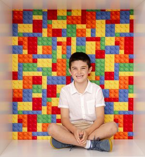 Smiling boy sitting cross-legged in front of a colorful LEGO brick backdrop in red, blue, yellow, and green.