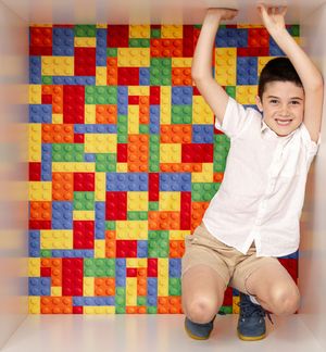A smiling boy in a white shirt crouches joyfully in front of a colorful LEGO brick wall backdrop.