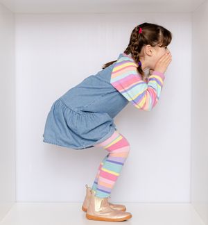 Young girl in striped top, denim dress, and gold boots crouching and covering her face with her hands.