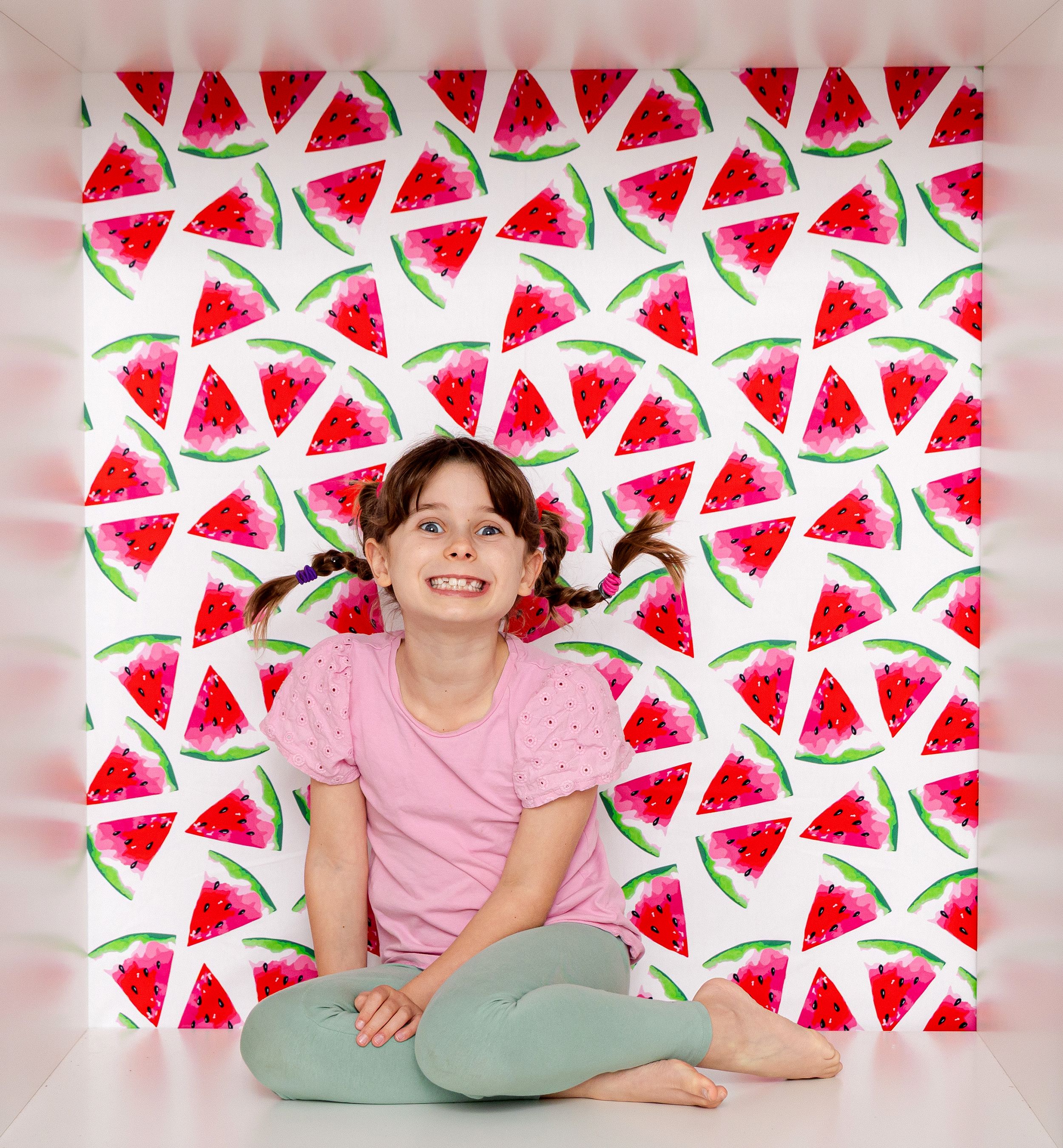 Smiling girl in pink shirt sits cross-legged against a colorful watermelon slice patterned backdrop.
