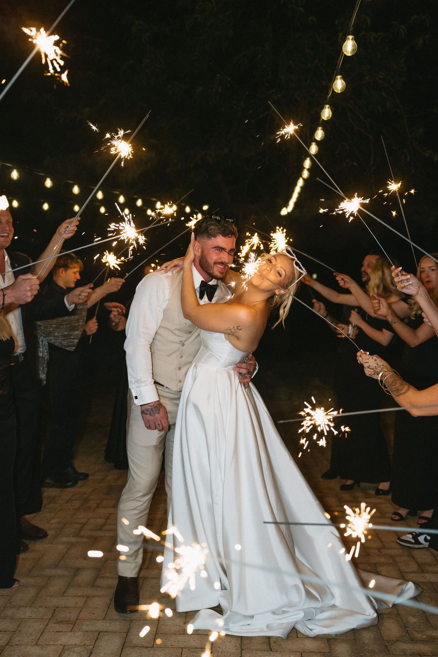 Bride and groom sharing a romantic kiss at sunset on a lush green lawn, wedding dress with long train glowing in golden light.