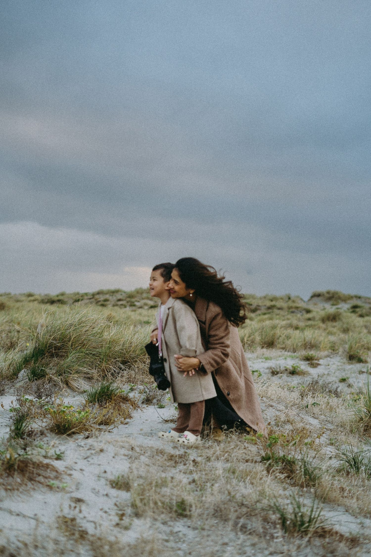 A woman with curly hair bends to kiss a young girl in a beige coat on a sandy coastal path with grassy dunes.