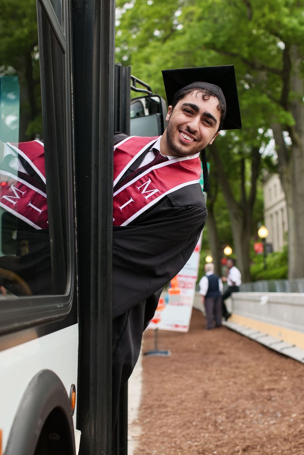 Smiling graduate in black gown and cap with red sash peers around a bus door on a tree-lined campus path.
