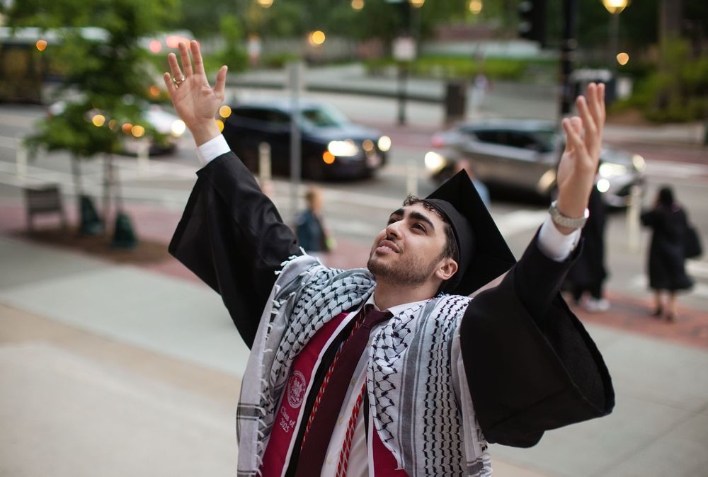 Graduate in cap and gown wearing keffiyeh raises hands skyward in joyful celebration outdoors.