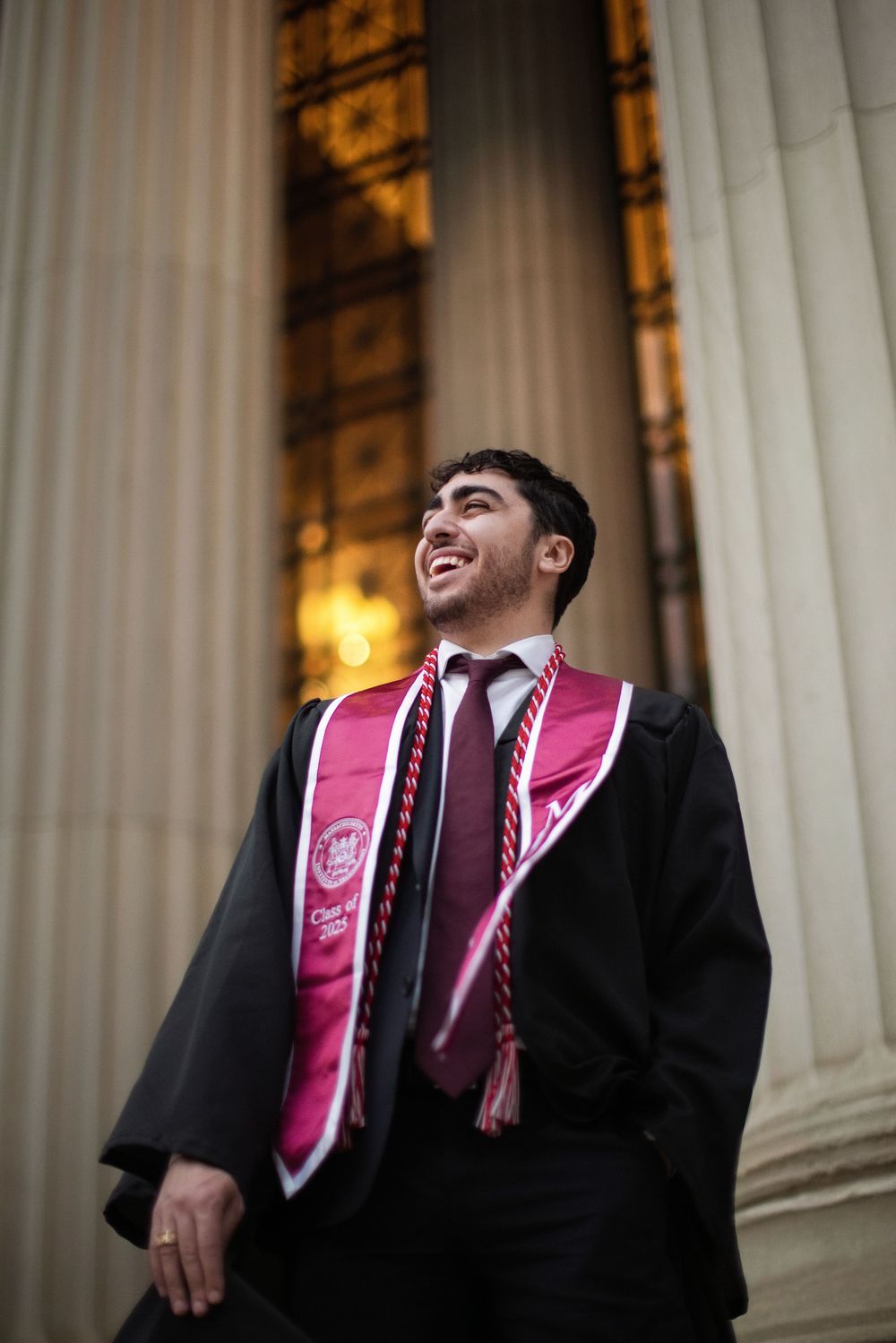 Smiling graduate in black gown and red sash stands before grand columns at commencement ceremony.