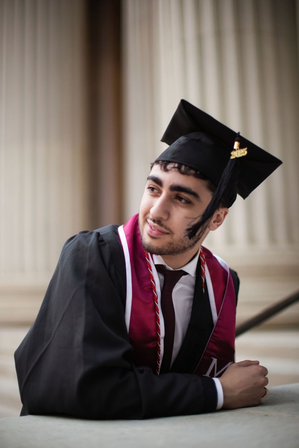 Young man in black graduation gown and cap with burgundy stole, smiling confidently while posing between columns.