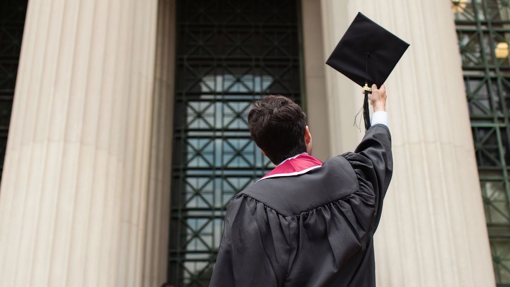 Graduate in black cap and gown raises mortarboard in front of a grand columned building.