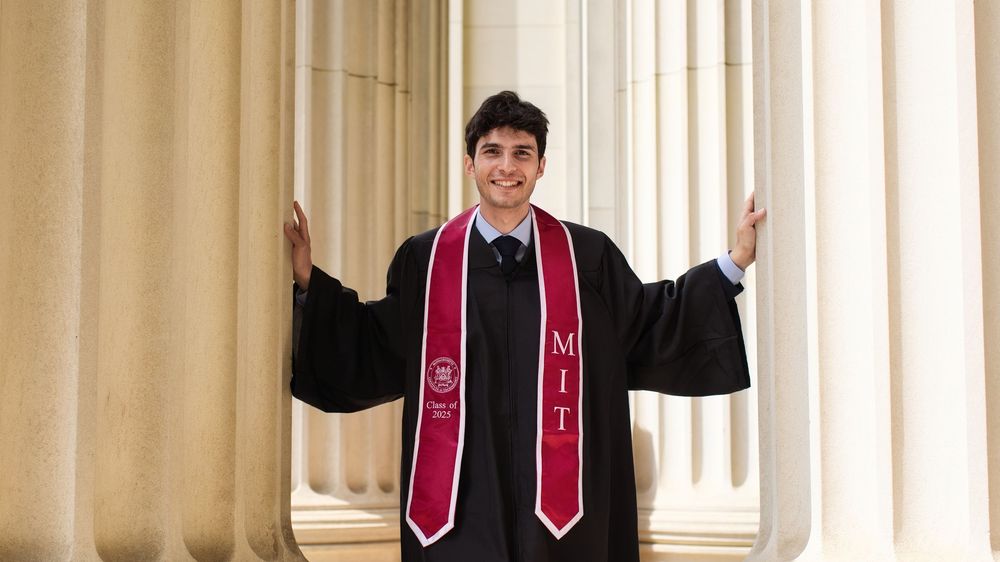 MIT graduate in black gown and red stole poses proudly between white columns at graduation ceremony.
