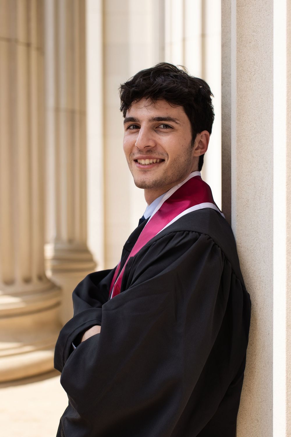 Smiling young man in black graduation gown with red stole leaning against a stone column.