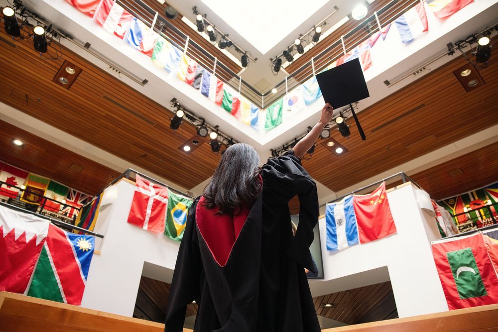 Graduate in cap and gown raises mortarboard in hall adorned with international flags during commencement ceremony.