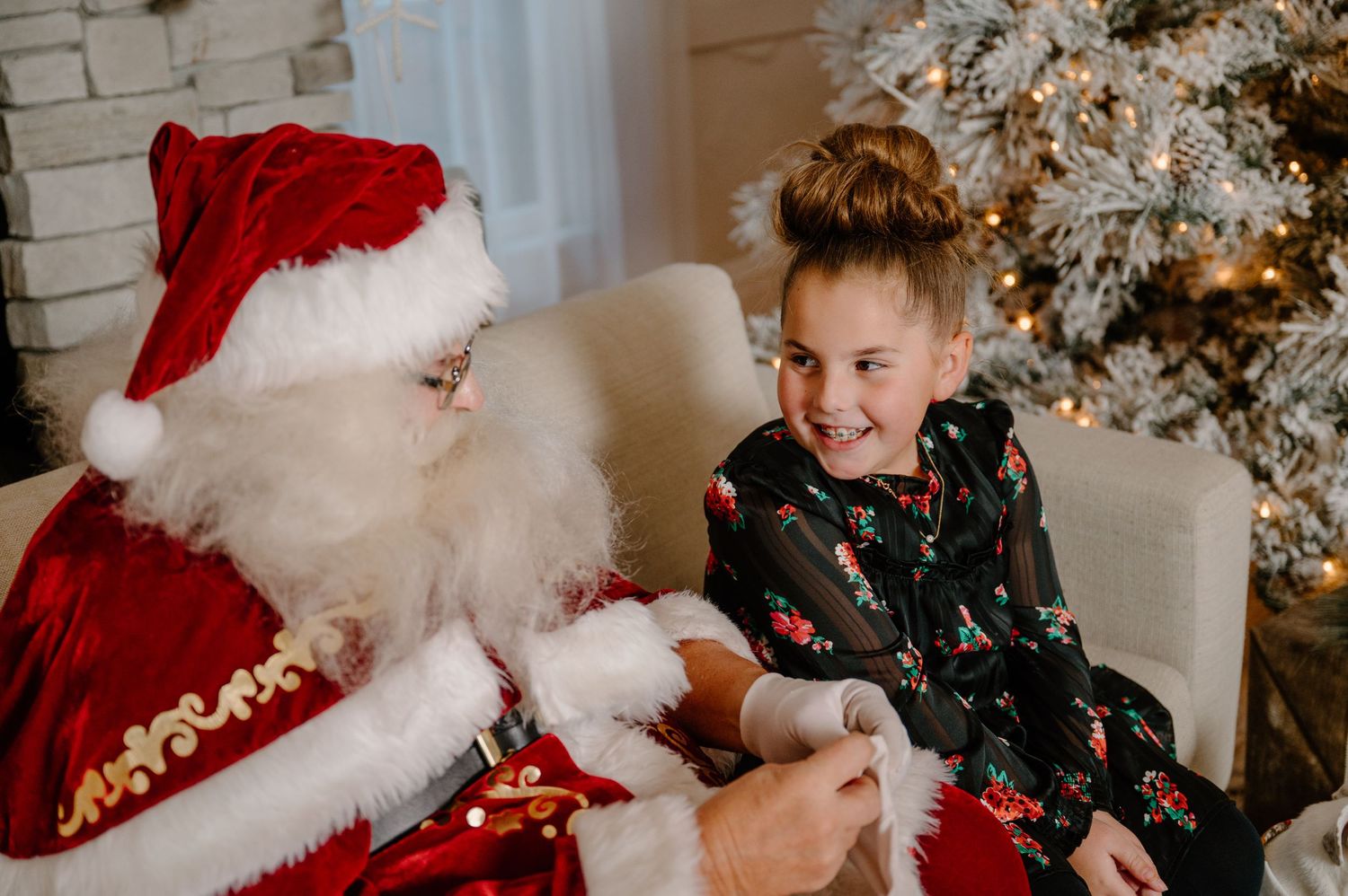 A joyful girl in a holiday dress laughs with Santa Claus beside a decorated Christmas tree.