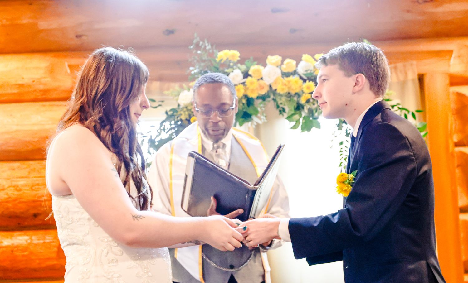 Couple exchanging vows at rustic log cabin wedding ceremony with officiant holding black book and yellow roses in background.