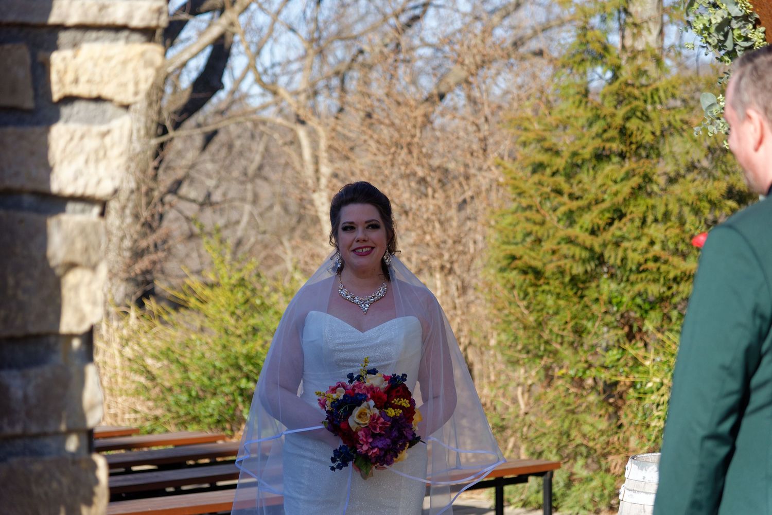 Bride in white gown and veil holding colorful bouquet smiling outdoors at wedding ceremony.