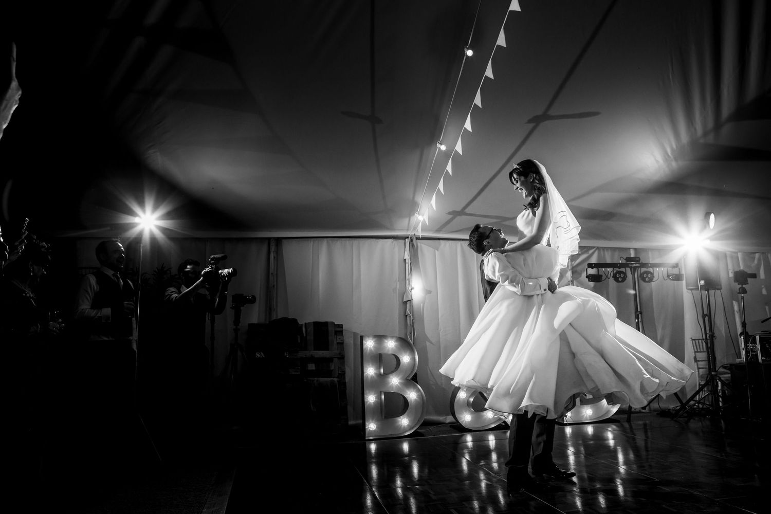Black and white photo of bride and groom sharing first dance at wedding reception with marquee lights.