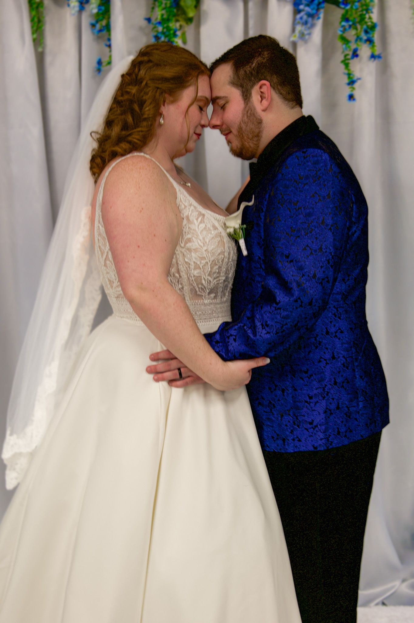 A bride and groom in wedding attire share an intimate moment, touching foreheads beneath a floral arch backdrop.