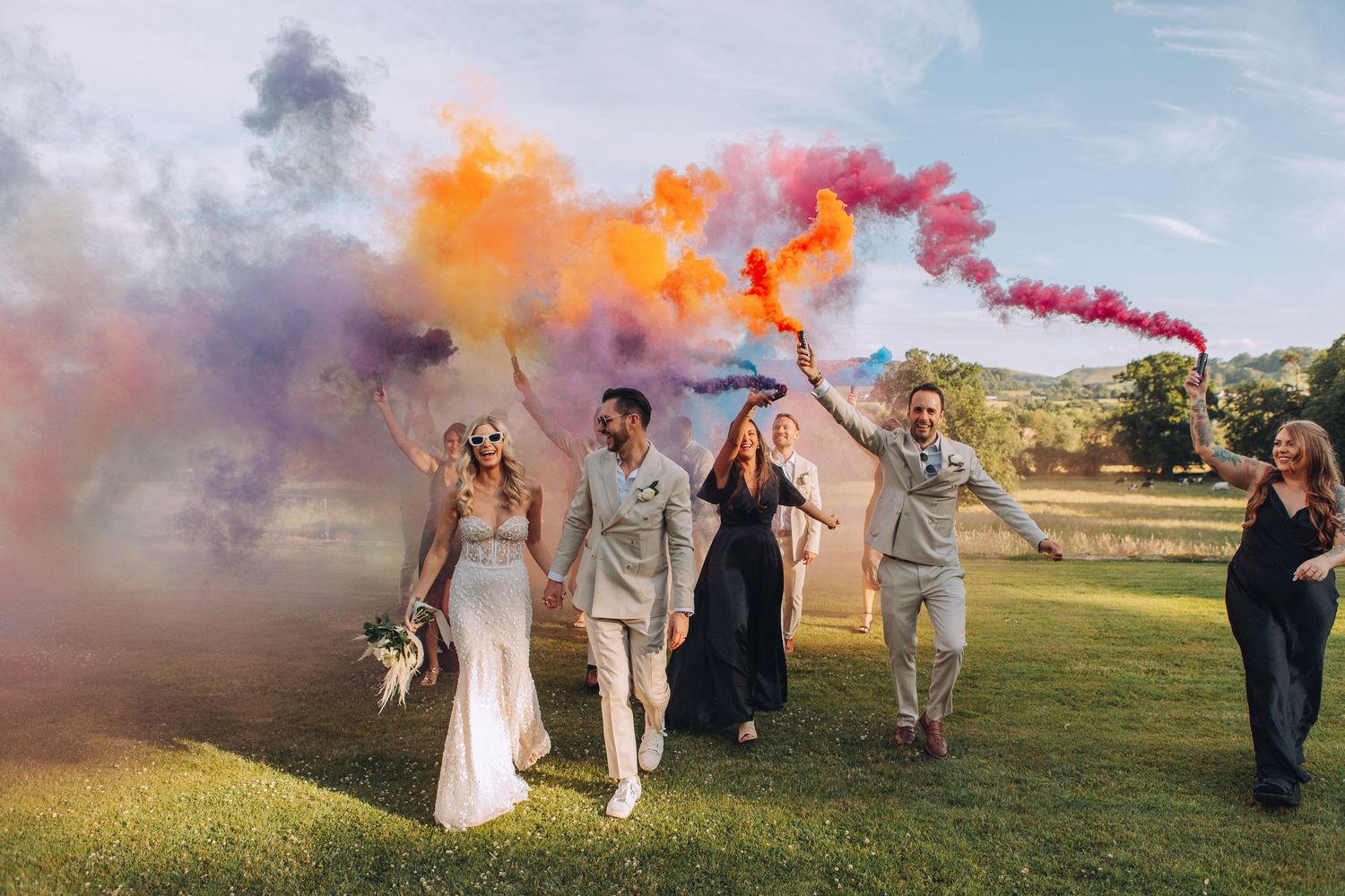 Wedding party celebrates outdoors with colorful smoke bombs in purple, orange, and red hues on a green lawn.