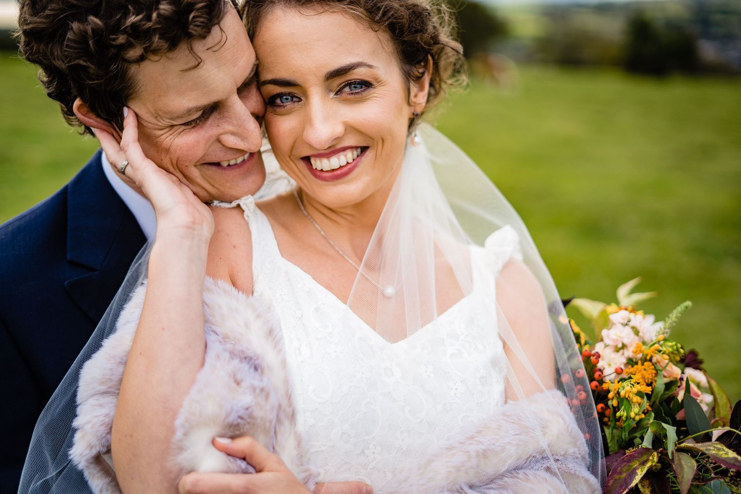 A joyful bride in a white gown and veil smiles warmly as her groom embraces her outdoors on their wedding day.