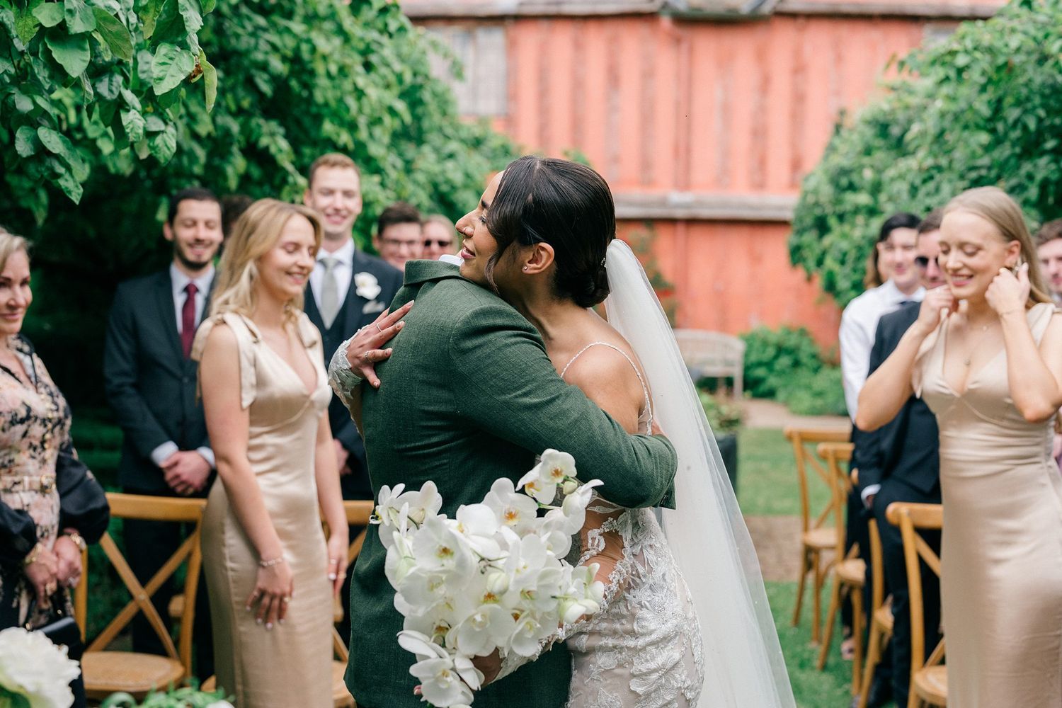 Bride and groom share a heartfelt hug at outdoor wedding ceremony with guests smiling in background.