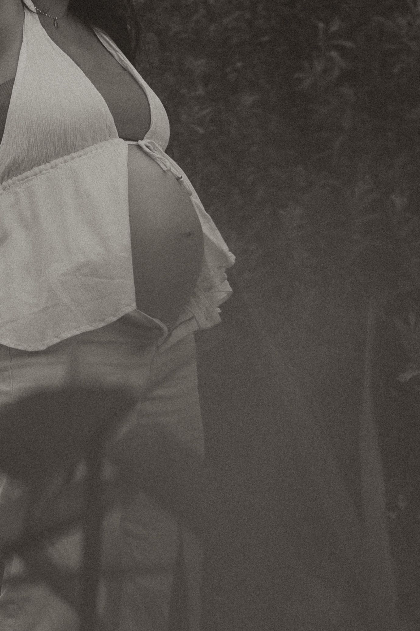 A couple sits on green grass, man in white tee kissing pregnant woman's belly, woman in denim jacket smiling softly.