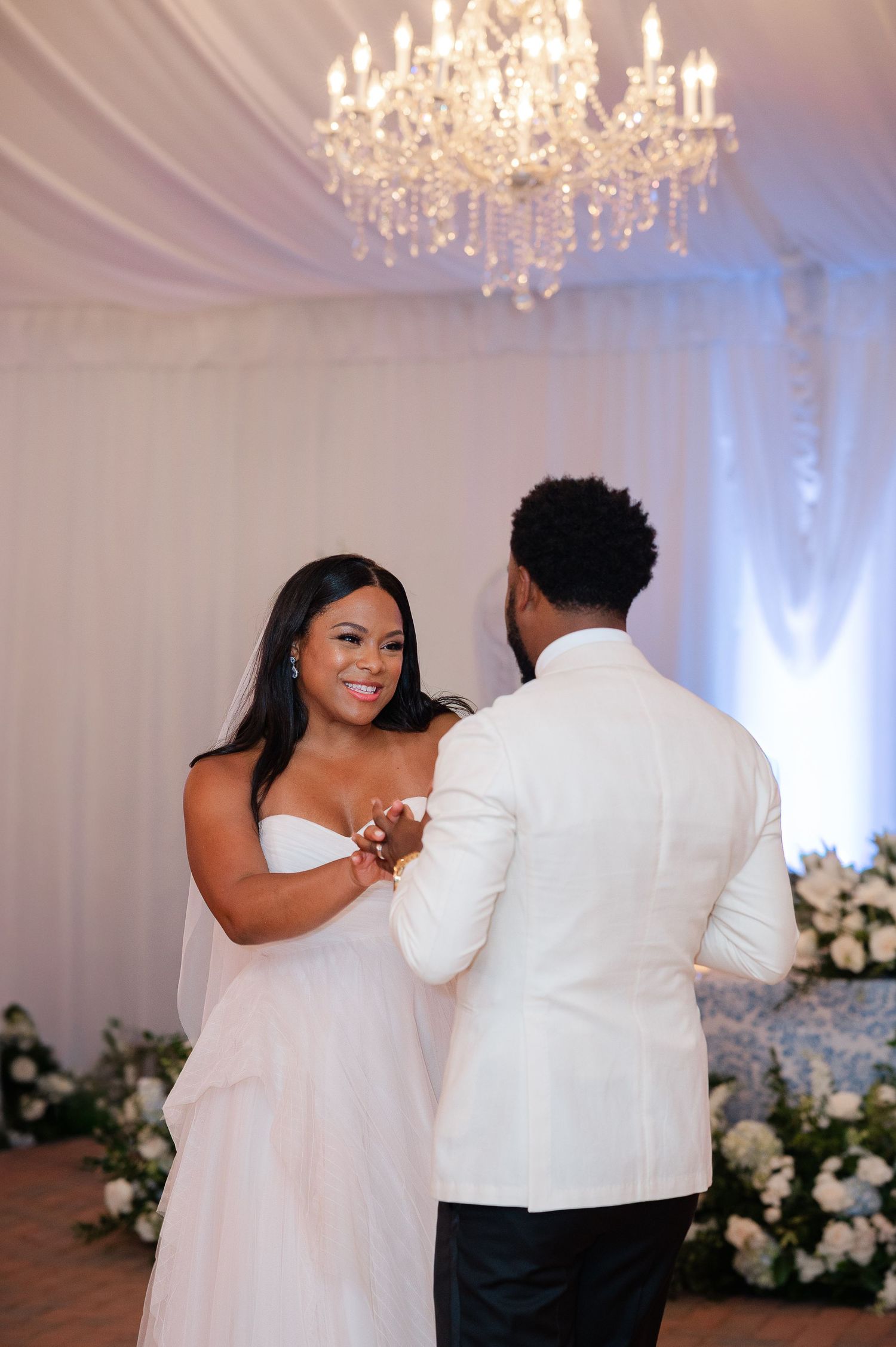 Bride and groom share their first dance under a crystal chandelier in an elegant white draped wedding reception venue.