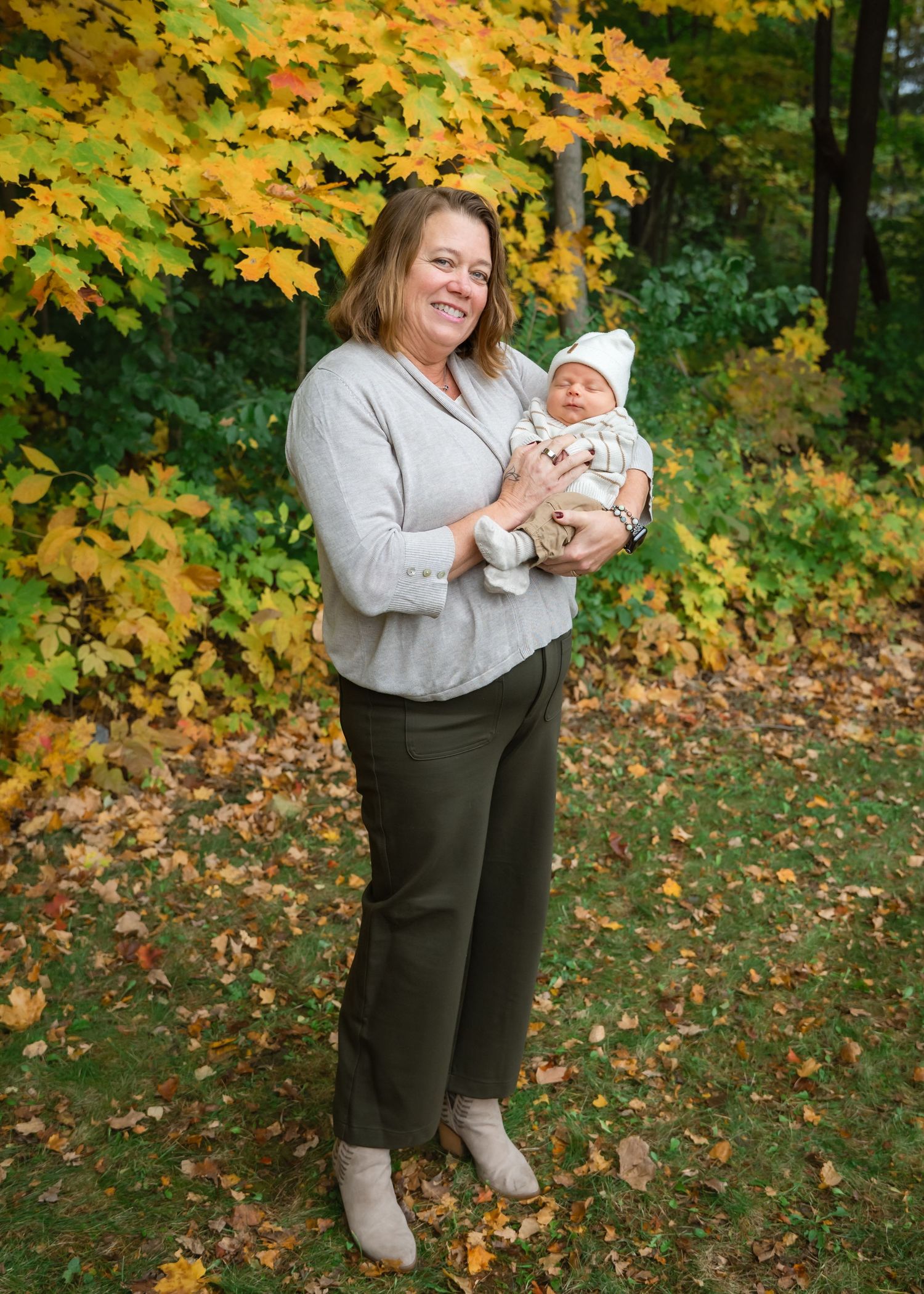 Woman smiling and holding a newborn baby outdoors during autumn with colorful fall foliage in the background.