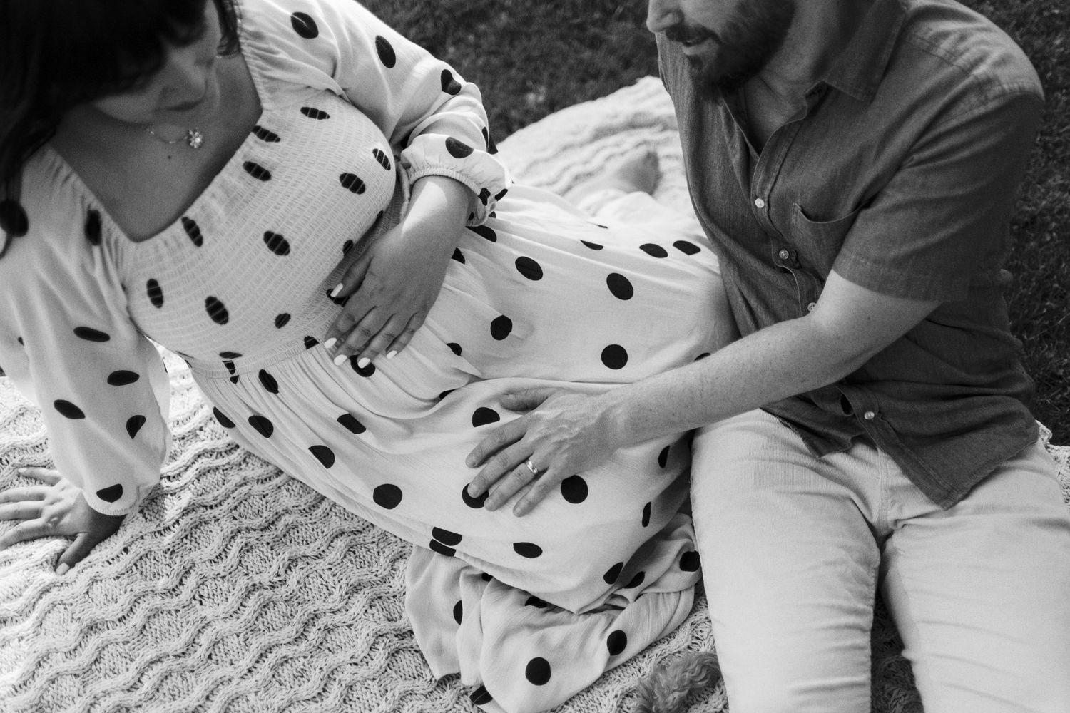 Black and white photo of a couple sitting outdoors, hands resting on pregnant belly in polka dot dress.