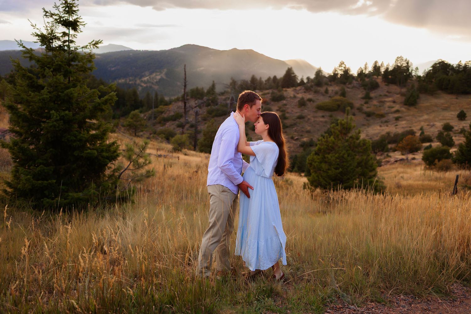 Couple sharing a romantic kiss in a golden mountain meadow at sunset, surrounded by pine trees and rolling hills.  Pregnant woman