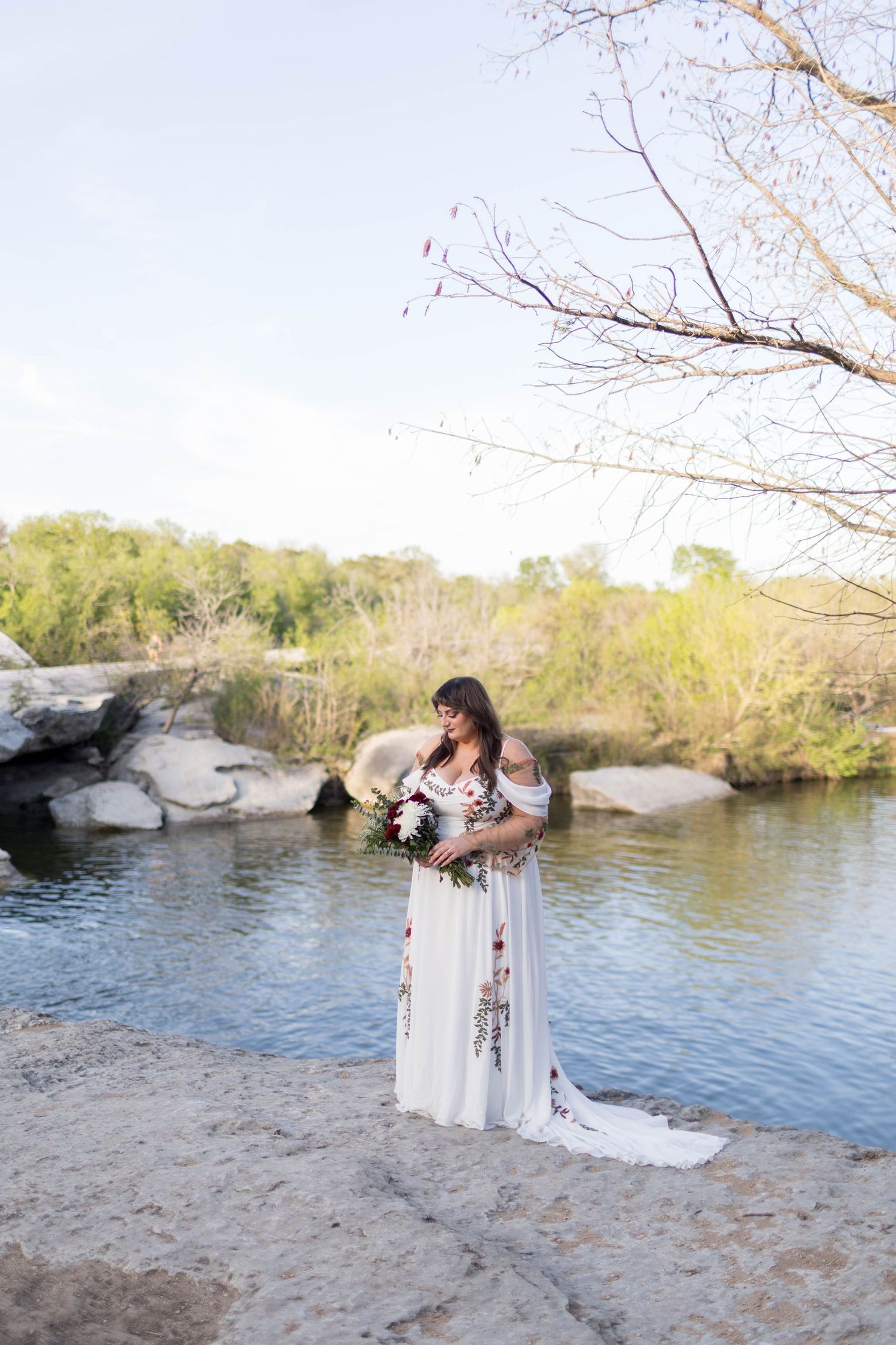 Bride in floral white gown holds bouquet by rocky riverside with lush green trees and clear sky backdrop.